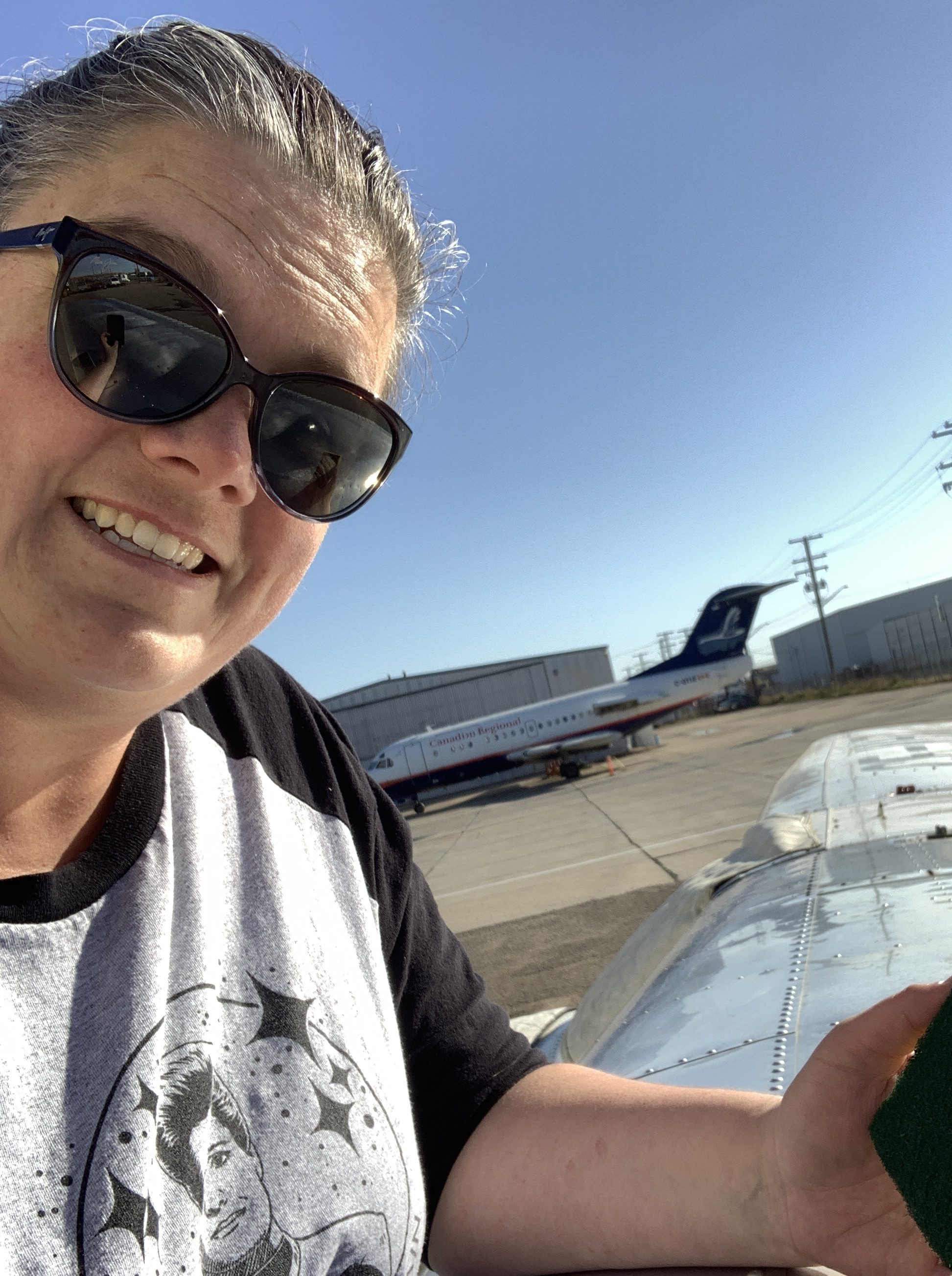 A picture of Desirae, a female with her hair pulled back, wearing sunglasses and a grey shirt with a black design on it and black sleeves. The person is leaning on the top of a high-wing airplane polishing the aluminum. The background is of a blue sky with a large commercial airplane in front of a storage hangar in the distance.